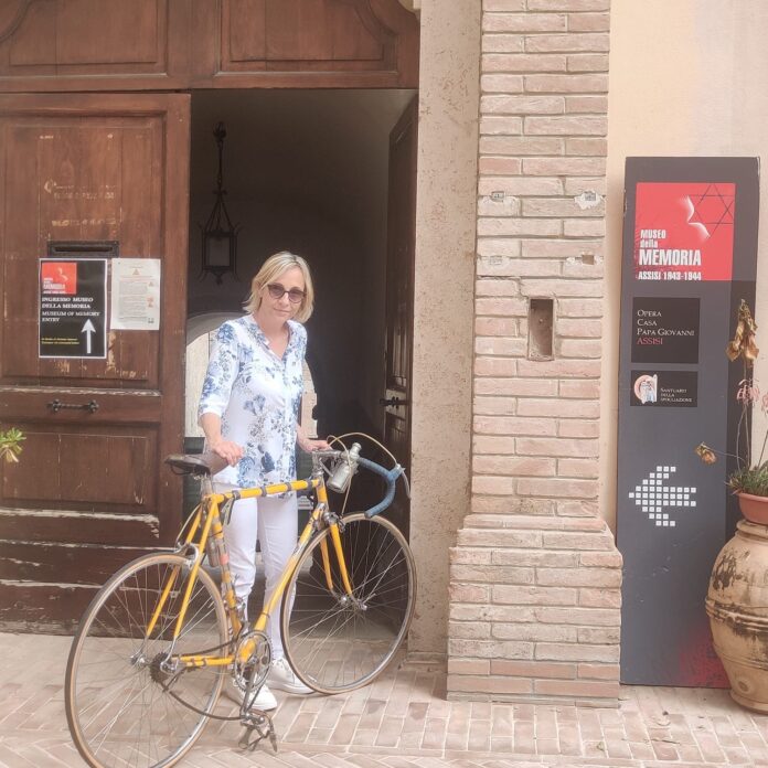 Gioia Bartali davanti alla porta di ingreso del Museo della Memoria di Assisi con la bicicletta del nonno Gino Bartali