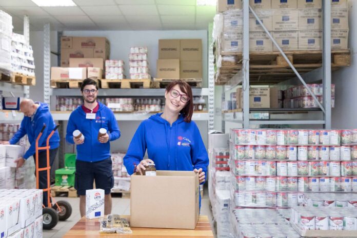 Un ragazzo e una ragazza con la maglietta blu del Servizio civile in primo piano, sullo sfondo un operatore adulto al lavoro in un magazzino di stoccaggio dei beni di prima necessità