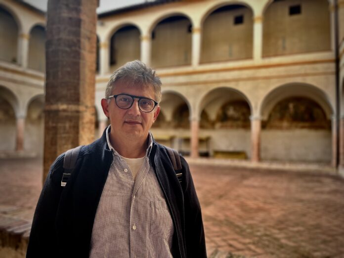 Mauro Masciotti a mezzo busto, con capelli grigi, gli occhiali, un giacchetto scuro e camichia celeste sullo sfondo un chiostro