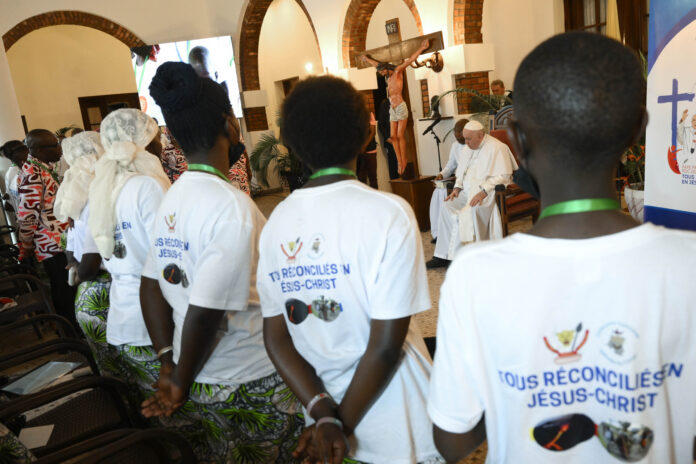 Uomini e donne congolosi di spalle con magliette bianche all'interno di una chiesa che guardano Papa Francesco seduto accanto ad un sacerdote congolese, vicino c'è una croce