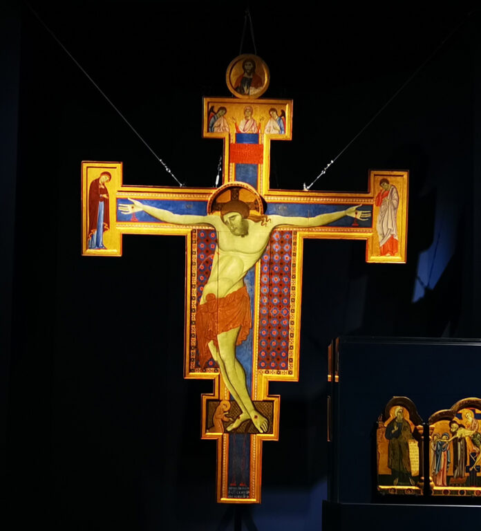 Croce monumentale in legnosostenuta da cavi, su fondo blu della sala espositiva, La croce è di colore blu profilata da conchiglie umbre, il tabellone centrale è decorato come un tappeto di persia. Al centro il Cristo con il corpo che pesa verso il basso, la testa e si incassa tra le spalle.Ha il perizoma rosso