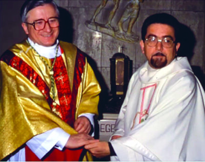 Don Orlando Sbicca a destra in veste liturgica bianca scringe la mano al vescovo Ennio Antonelli, a sinistra, in veste liturgica color oro e rosso.