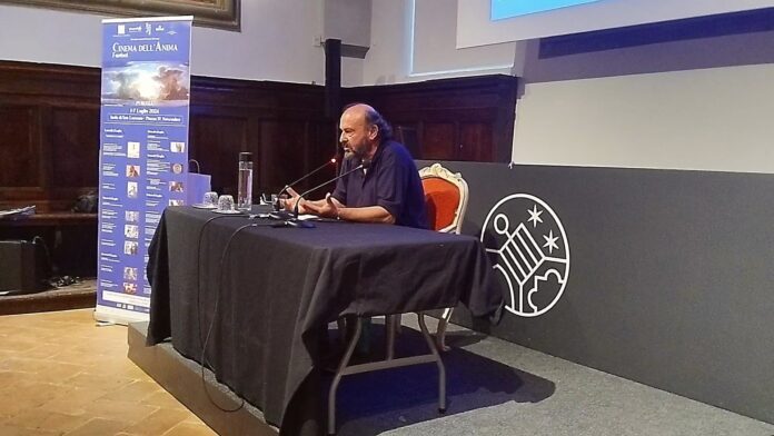 Davide Rondoni in maglietta blu, seduto ad un tavolo, di profilo, sullo sfondo la locandina del festival “Cinema dell'anima