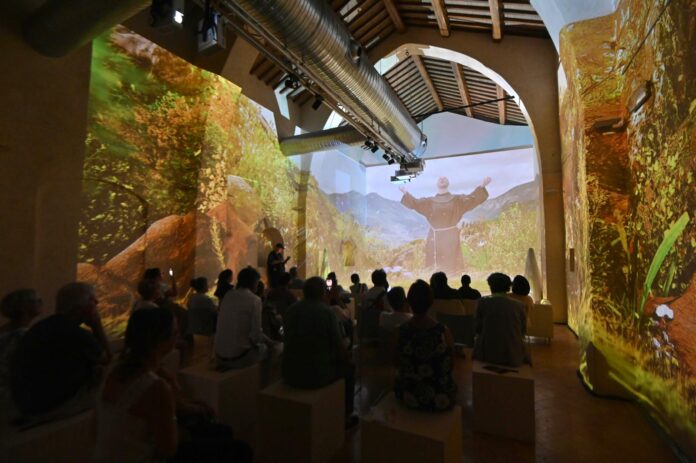 Sullo sfondo le parte delle stanza dove è prioiettata l'immaginbe di fra Brustegnhi che canta a braccia aperte, nei due schermi laterali immagini di natura. in primo piano teste di spalle di persone presente scure