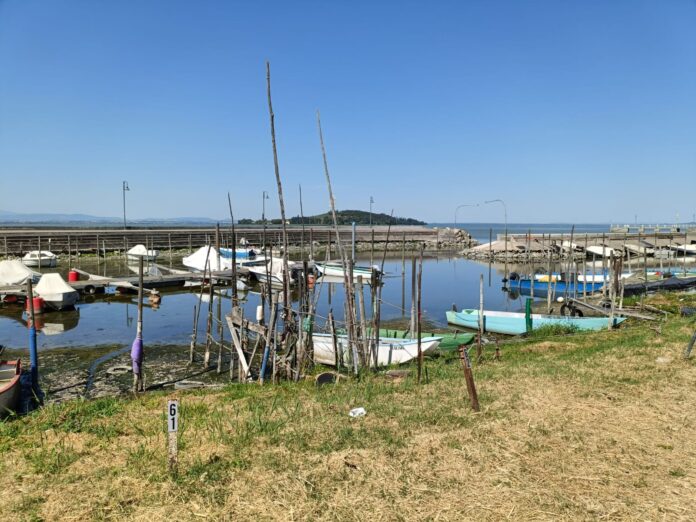 In primo piano la sabbia della costa del lago Trasimeno e sullo sfondo il lago con i canneti e le barche e dietro la costa dell'altra spiaggia