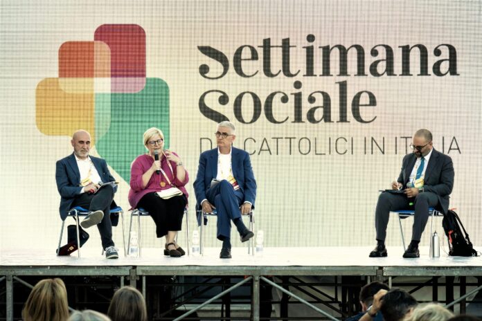 Quattro relatori , di cui una donna, seduti sulle sedie sul palco. Sullo sfondo la locandina della Settimana sociale