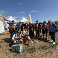 Un gruppo di capi scout umbri con la camicia azzurra e il fazzolettone in piedi e due in primo piano inginocchiato e seduto con in mano la chitarra, sullo sfondo le tensostrutture bianche