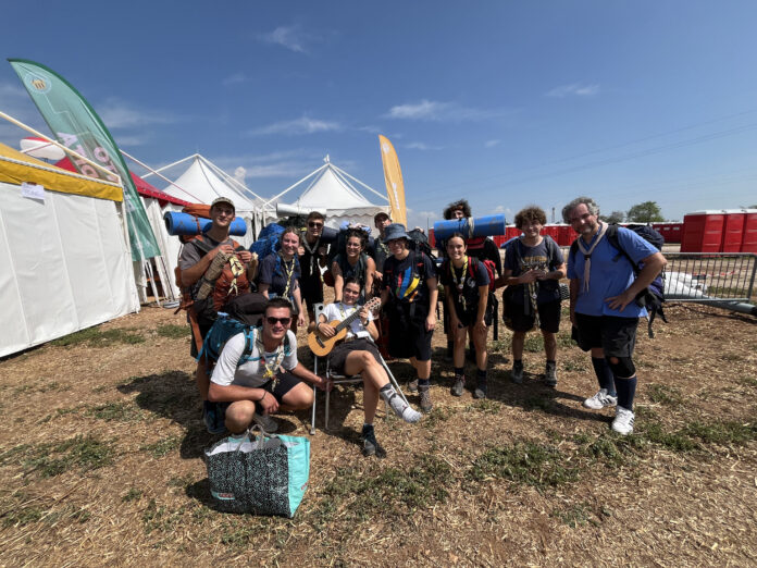 Un gruppo di capi scout umbri con la camicia azzurra e il fazzolettone in piedi e due in primo piano inginocchiato e seduto con in mano la chitarra, sullo sfondo le tensostrutture bianche