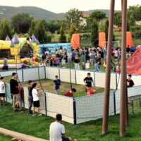 Bambini e ragazzi che giocano a pallone all'interno di un piccolo campo da gioco dal recinto bianco, all'interno di un'area verde, iall'esterno dell'area da gioco altri ragazzi che guardano e intorno tra il verde altri giocano con i gonfiabili