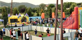 Bambini e ragazzi che giocano a pallone all'interno di un piccolo campo da gioco dal recinto bianco, all'interno di un'area verde, iall'esterno dell'area da gioco altri ragazzi che guardano e intorno tra il verde altri giocano con i gonfiabili