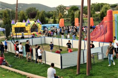 Bambini e ragazzi che giocano a pallone all'interno di un piccolo campo da gioco dal recinto bianco, all'interno di un'area verde, iall'esterno dell'area da gioco altri ragazzi che guardano e intorno tra il verde altri giocano con i gonfiabili