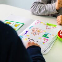 Le braccia di un bambino che guarda davanti a sè un foglio con dei disegni, di fronte alla maestra che è di spalle, con un maglia nera, e di cui si vede solo la spalla e il braccio destro e la mano