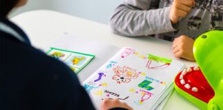 Le braccia di un bambino che guarda davanti a sè un foglio con dei disegni, di fronte alla maestra che è di spalle, con un maglia nera, e di cui si vede solo la spalla e il braccio destro e la mano