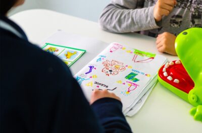 Le braccia di un bambino che guarda davanti a sè un foglio con dei disegni, di fronte alla maestra che è di spalle, con un maglia nera, e di cui si vede solo la spalla e il braccio destro e la mano