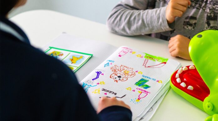 Le braccia di un bambino che guarda davanti a sè un foglio con dei disegni, di fronte alla maestra che è di spalle, con un maglia nera, e di cui si vede solo la spalla e il braccio destro e la mano