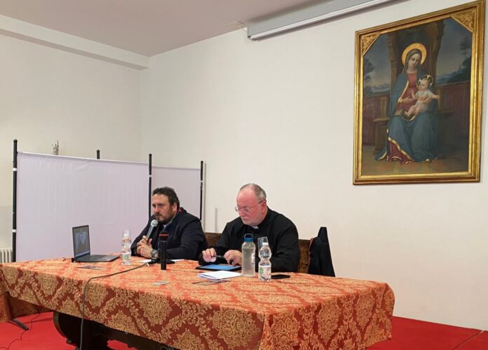 Don Francesco Verzini a sinistra e a destra don Fabio Rosini seduti dietro ad un tavolo, visti di tre quarti e alle spalle un quadro con ritratto la Madonna con il Bambino