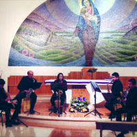 In primo piano il quartetto di musicisti seduti che suonano lo strumento a fiato, alle loro spalle l'altare della cappella dell'ospedale con sullo sfondo un'immagine della Madonna con il Bambino affrescata sul muro bianco che ritrae la Madonna con il bambino sullo sfondo una mezzaluna che rappresenta colline di vari colori