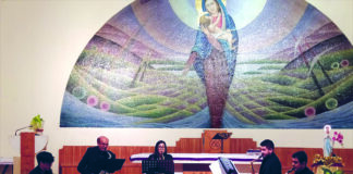 In primo piano il quartetto di musicisti seduti che suonano lo strumento a fiato, alle loro spalle l'altare della cappella dell'ospedale con sullo sfondo un'immagine della Madonna con il Bambino affrescata sul muro bianco che ritrae la Madonna con il bambino sullo sfondo una mezzaluna che rappresenta colline di vari colori