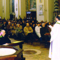 Un sacerdote in piedi di spalle di tre quarti in chiesa vicino all'altare, davanti a lui i fedeli seduti nelle panche