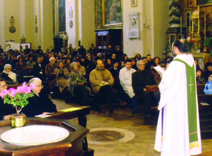 Un sacerdote in piedi di spalle di tre quarti in chiesa vicino all'altare, davanti a lui i fedeli seduti nelle panche