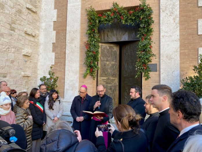 Il vescovo Ivan Maffeis. con il vicario all'apertura dell'anno santo, alle loro spalle la porta santa della facciata laterale della cattedrale circondata da racemi vegetali. Davanti alcune persone e autorità