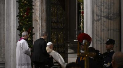 Papa Francesco seduto sulla sedia a rotelle davanti alla porta santa aperta di San Pietro il giorno dell'apertura, accompagnato da un sacerdote