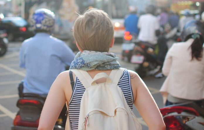 gente di spalle, qualcuno con il motorino, in primo piano una donna con i capelli biondi e corti con canottiera, foulard intorno al collo e zaino in spalla