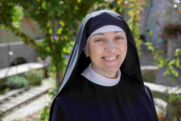 Suor Maria Grazia sorridente ritratta a mezzo busto, con l'abito monacale e il velo nero, sullo sfondo rami di alberi