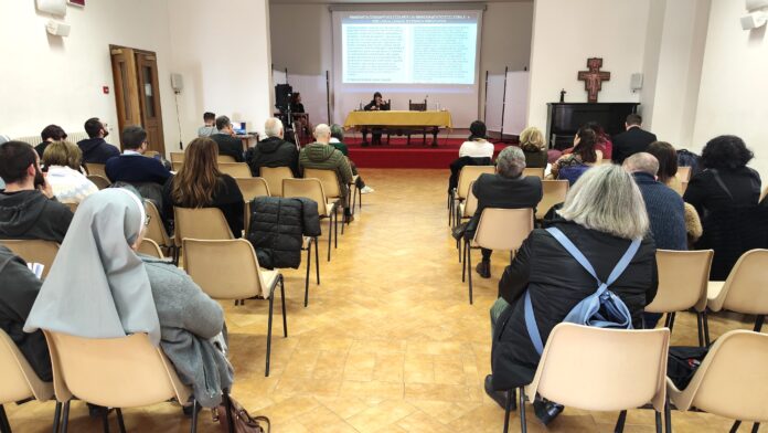 Gente del pubblico di spalle seduti e in fondo il tacolo dei relatori del convegno all'interno di una sala del seminario di Assisi